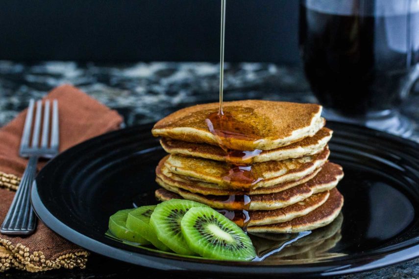 Maple syrup being poured over 3 Seed dairy-free gluten-free Pancakes on a plate served with sliced kiwi. 