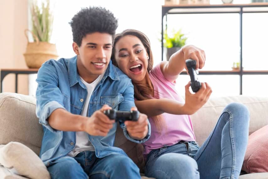 Happy black teenagers playing video game with control pad, holding joysticks in living room