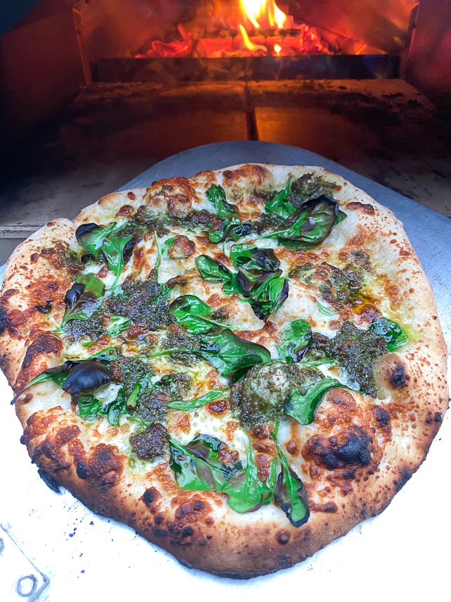 pesto spinach pizza on peel in front of wood fired Ooni oven