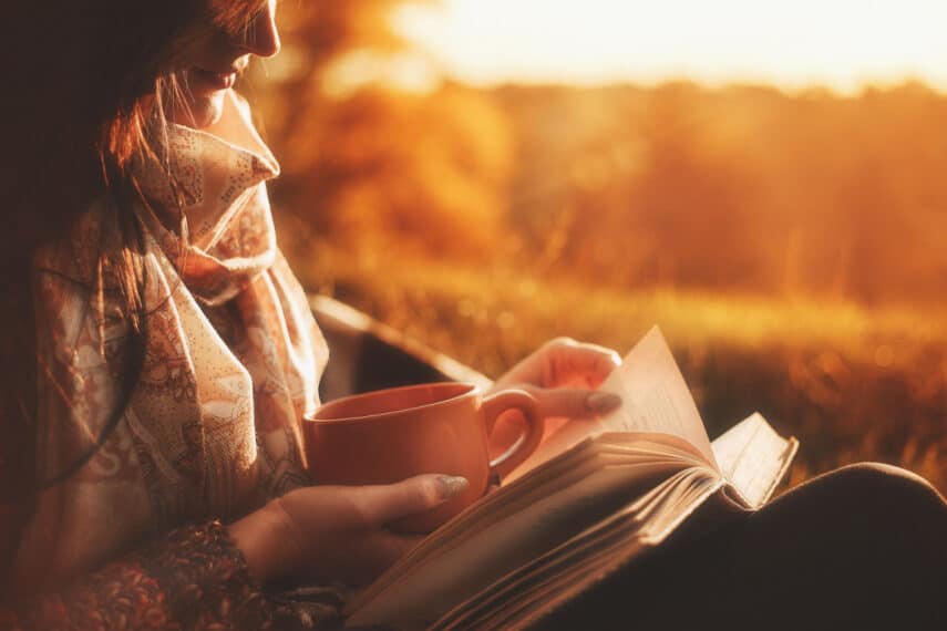 Beautiful girl in autumn forest reading a book covered with a warm blanket.a woman sits near a tree in an autumn forest and holds a book and a cup with a hot drink in her hands. Girl reading a book.