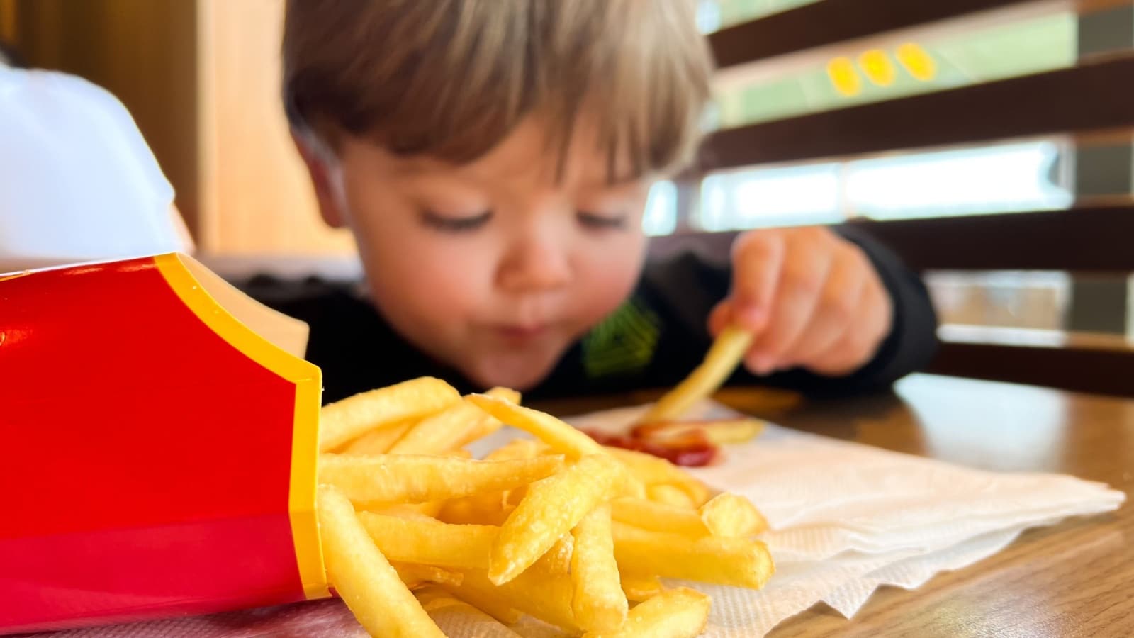 A little boy, caucasian child eating McDonald's fries with ketchup inside McDonald's restaurant. Unhealthy fast food eaten by a kid.