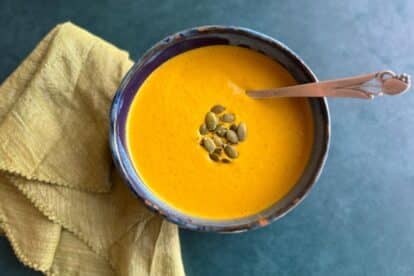 Low FODMAP Roasted Carrot Tahini Soup in a ceramic bowl on teal background with spoon inserted.