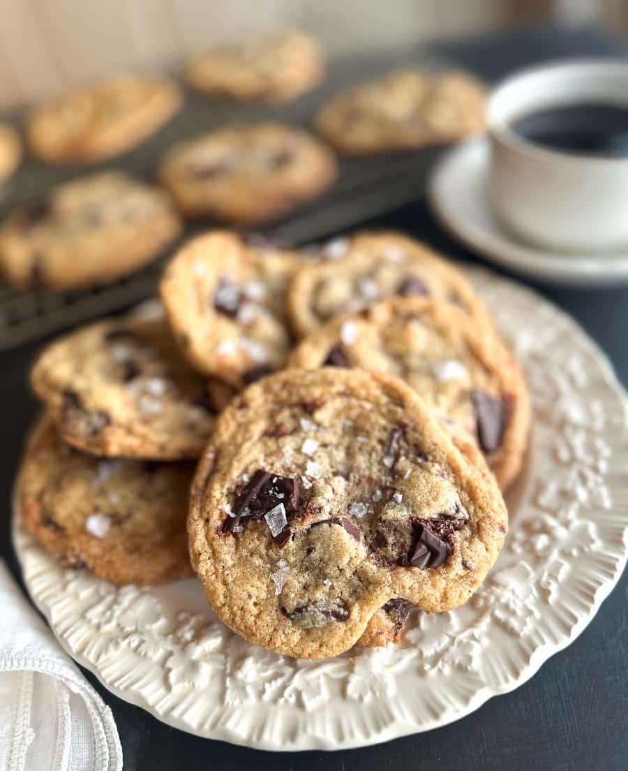 Vertical Low FODMAP Tahini Chocolate Chunk Cookies on white plate and rack. Cup of coffee.