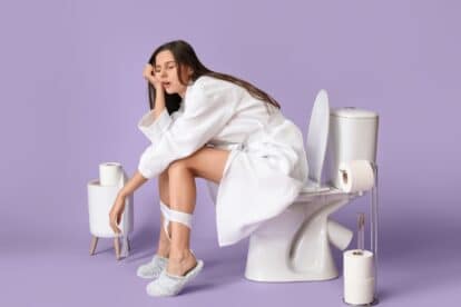 Sleepy young woman in bathrobe sitting on toilet bowl against color background