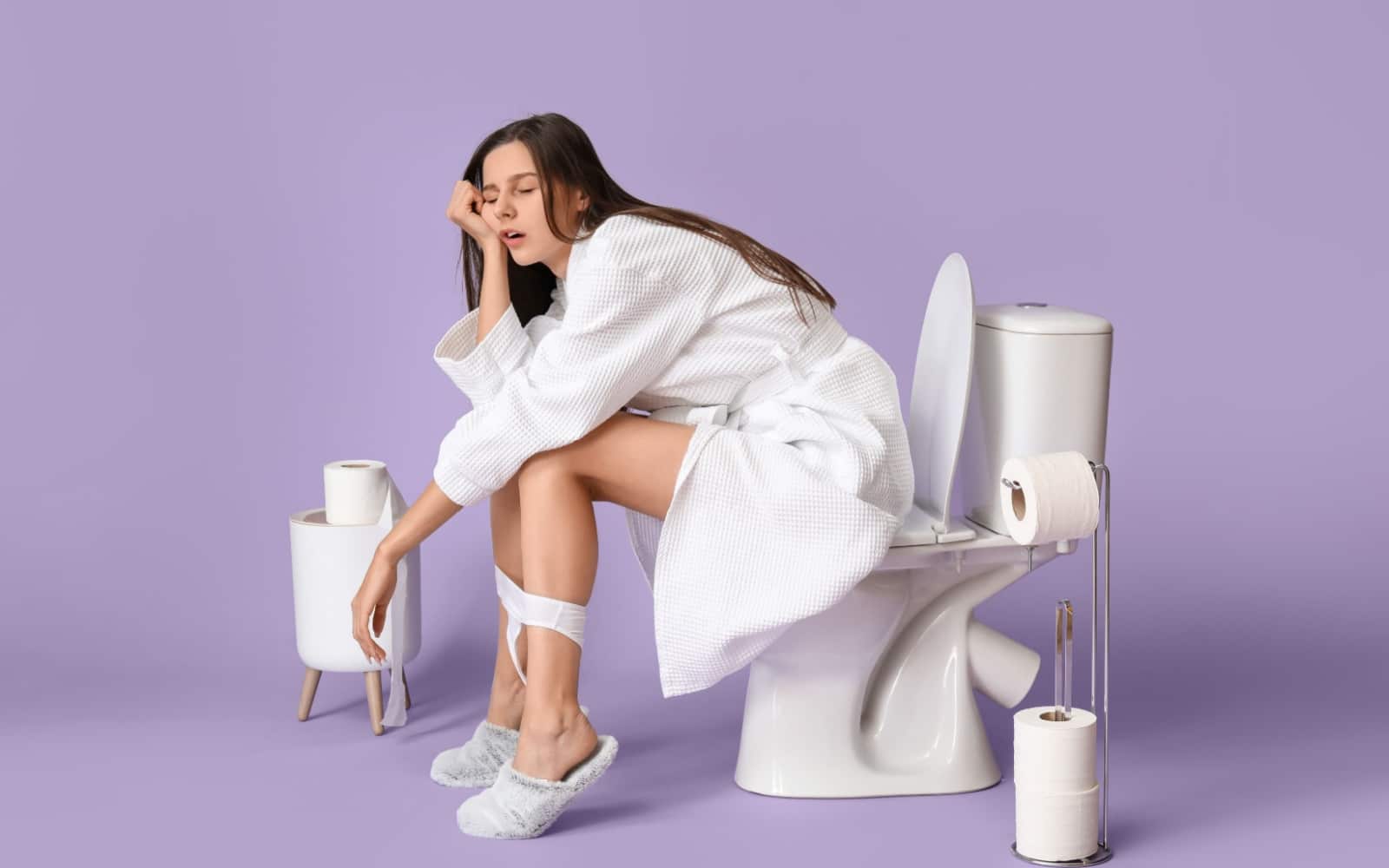 Sleepy young woman in bathrobe sitting on toilet bowl against color background